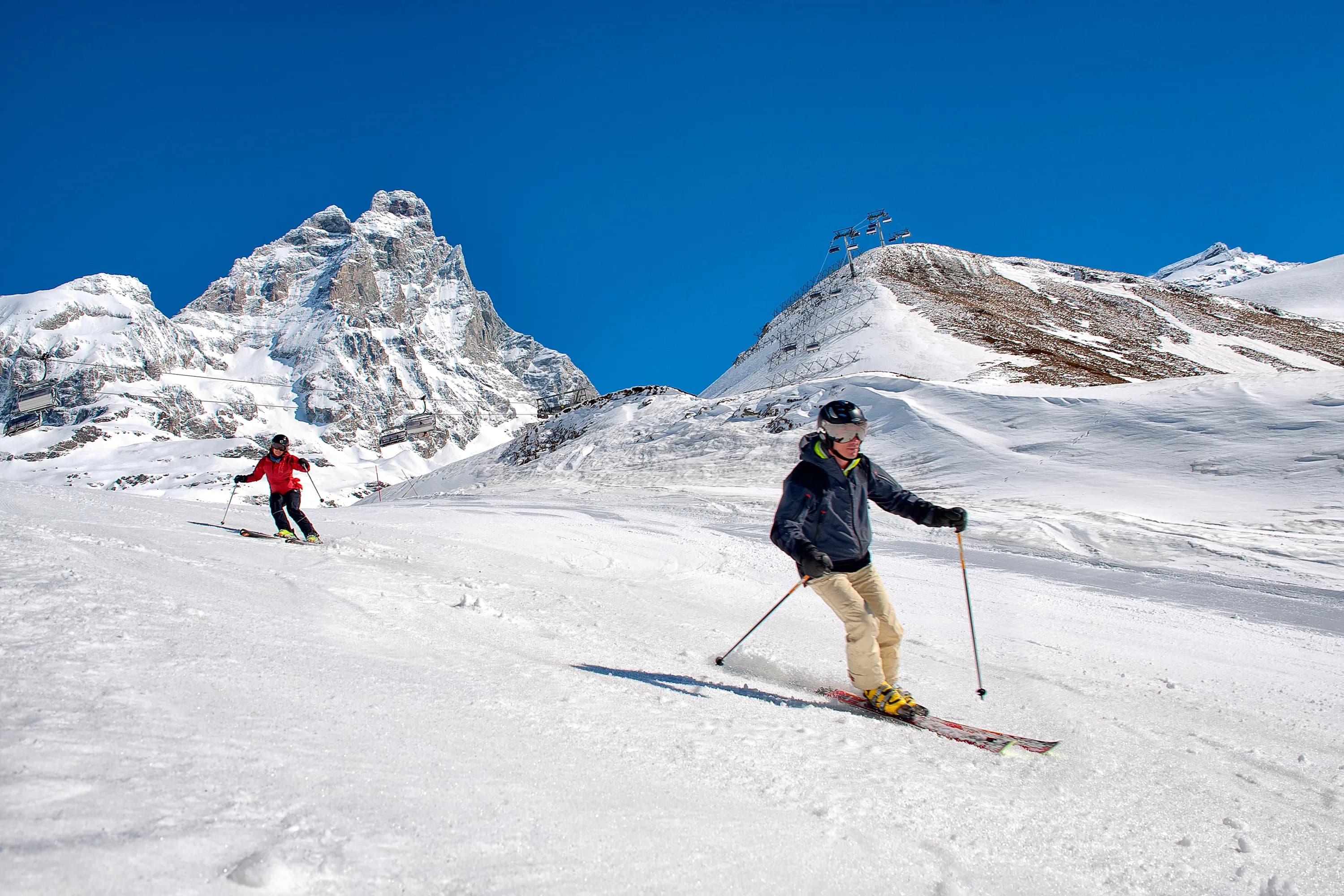 Två skidåkare susar nerför backen vid Cervinia, med det majestätiska Matterhorn i bakgrunden och omgivna av snötäckta alper.